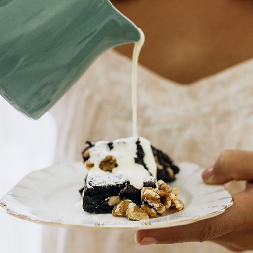 A person is pouring cream on a brownie with nuts, served on a white plate.