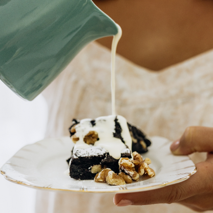 A person is pouring cream on a brownie with nuts, served on a white plate.
